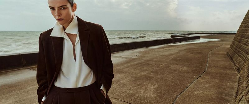 Fashion shot of a woman with short dark hair wearing a brown suit with a white blouse posing outdoors with the ocean in the background.