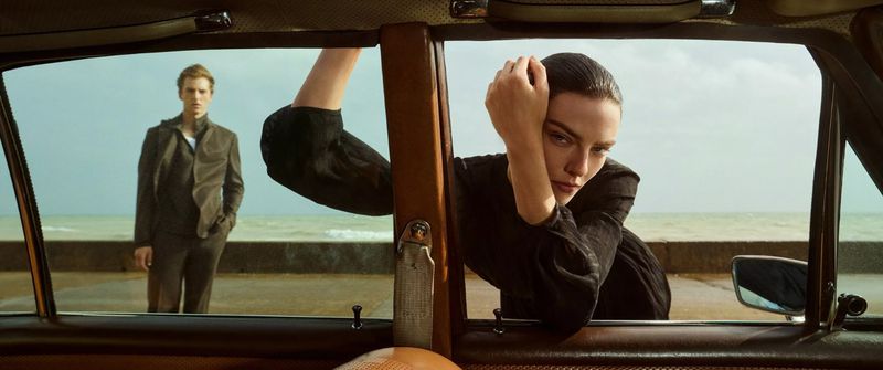 Indoor shot through car window of woman in black leaning on door and man standing on the beach in the background, ocean view.