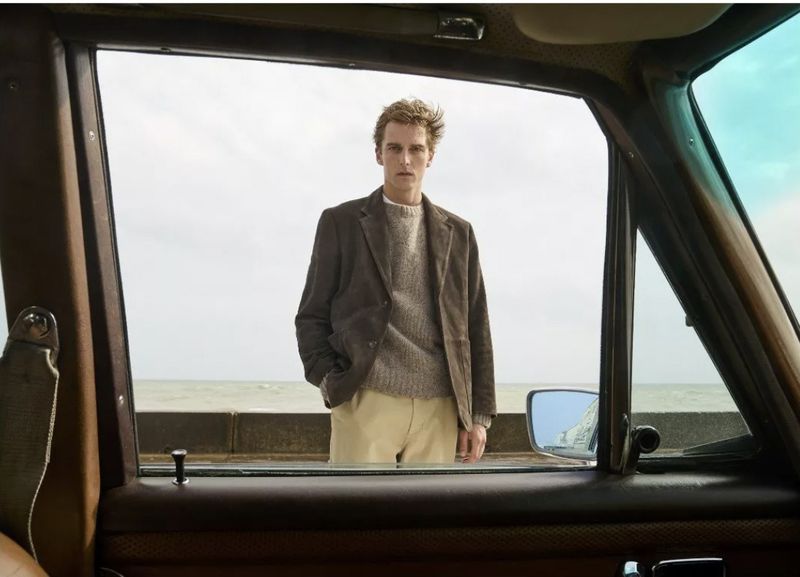 A stylish adult male in a brown jacket and beige pants stands in front of a sea wall, framed by an open car window. Ocean backdrop.