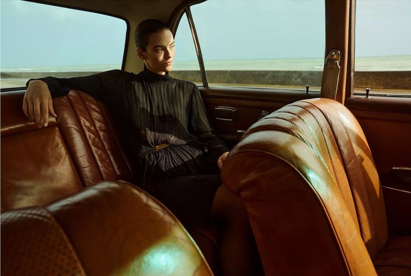 Adult woman in a black dress is sitting on a brown leather car seat, looking out the window at a blue sea and sky in natural lighting.