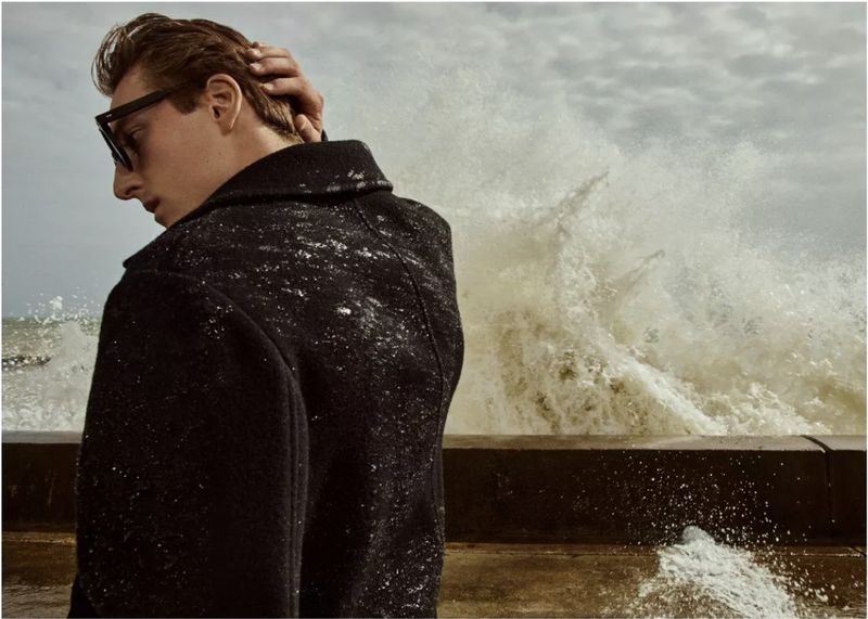 Adult male model in black coat and sunglasses with hand to head, photographed on a windy day with waves crashing in the background.