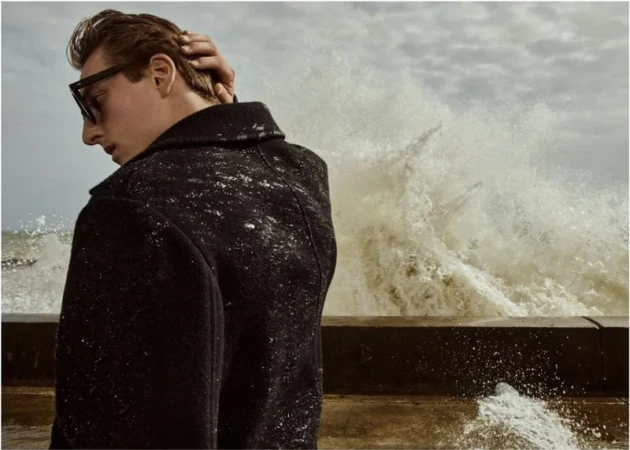 Adult male model in black coat and sunglasses with hand to head, photographed on a windy day with waves crashing in the background.