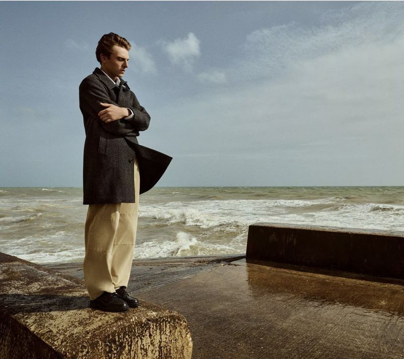 Young man in a coat stands on a concrete structure near the sea with a cloudy sky in the background, looking thoughtfully towards the water.