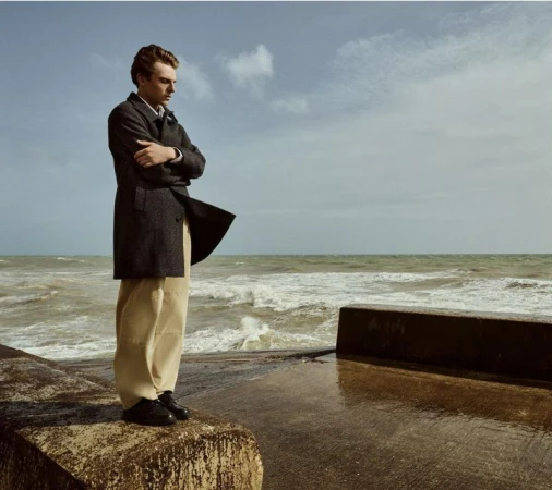 Young man in a coat stands on a concrete structure near the sea with a cloudy sky in the background, looking thoughtfully towards the water.