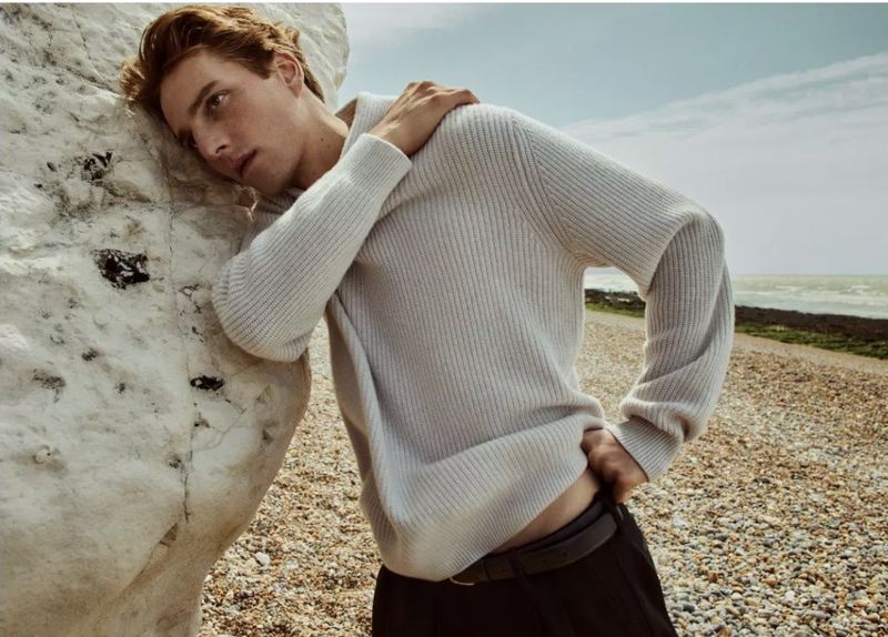 Quentin Demeester wears an off-white ribbed knit sweater leaning against a chalk cliff on a pebbled beach for Vince F/W 2025.