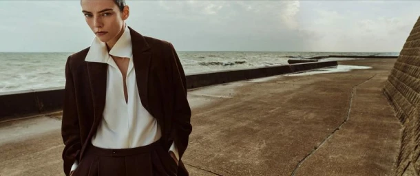 Model Beauïse Genç wearing a dark brown tailored suit and open-collar cream blouse standing beside a concrete seawall by the ocean.