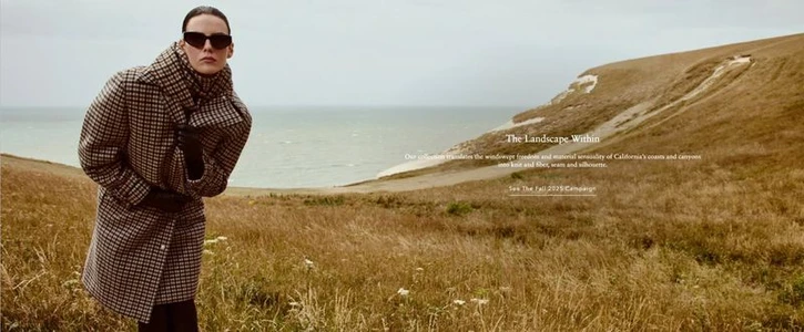 Model wearing a plaid coat and dark sunglasses standing in a coastal grassy field above the ocean.