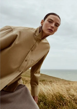 Model Beauïse Genç leaning forward in a beige high-collar button-up shirt against a coastal grassy field and ocean.