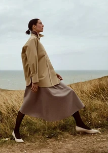 Model posing in an oversized beige collared shirt and taupe midi skirt on a grassy coastal bluff overlooking the ocean.