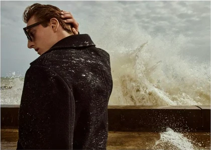 Male model in a dark textured coat wearing sunglasses by a crashing ocean wave for Vince F/W 2025 Campaign