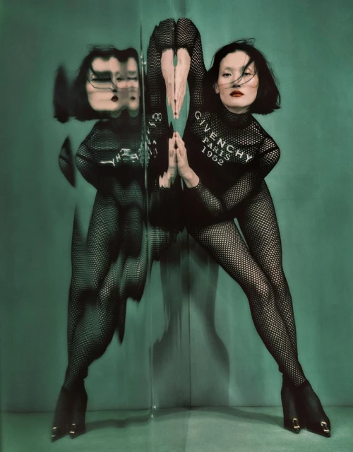 Woman in fishnet bodysuit poses with a mirror reflecting her image, set against a green backdrop; fashion-forward and artistic composition.