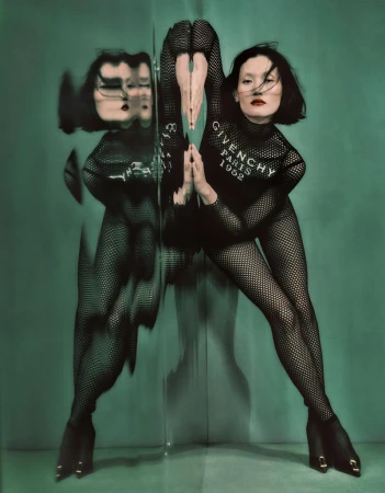 Woman in fishnet bodysuit poses with a mirror reflecting her image, set against a green backdrop; fashion-forward and artistic composition.