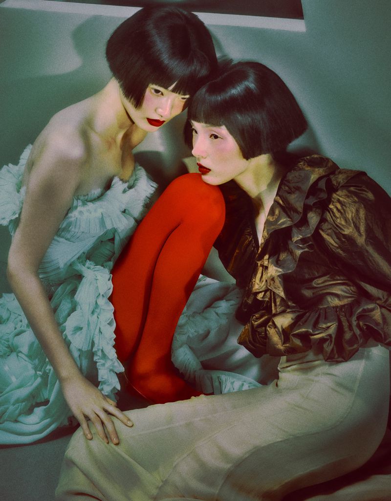 Two Asian women pose in avant-garde fashion, wearing pale dresses and red lipstick, with matching bob haircuts indoors.