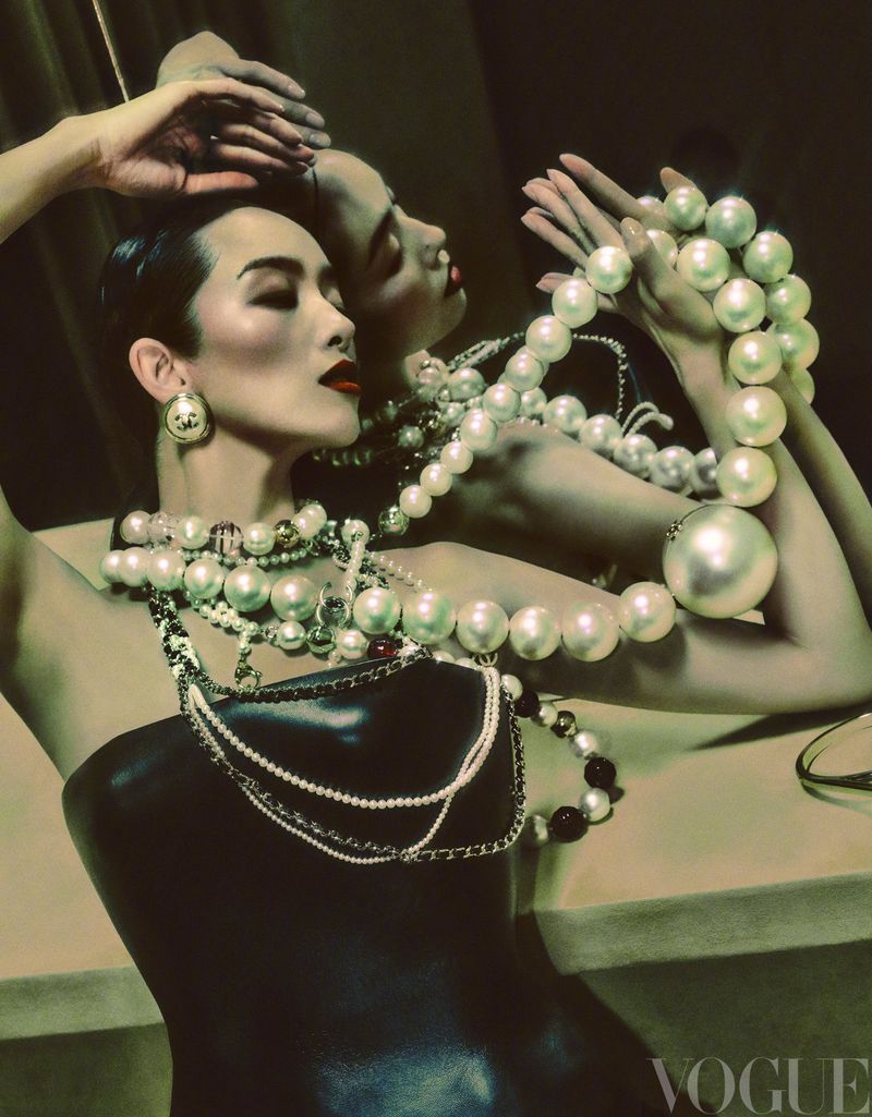 Glamour portrait of a woman wearing a black top and layers of pearl necklaces, with red lipstick and her hands near her face.