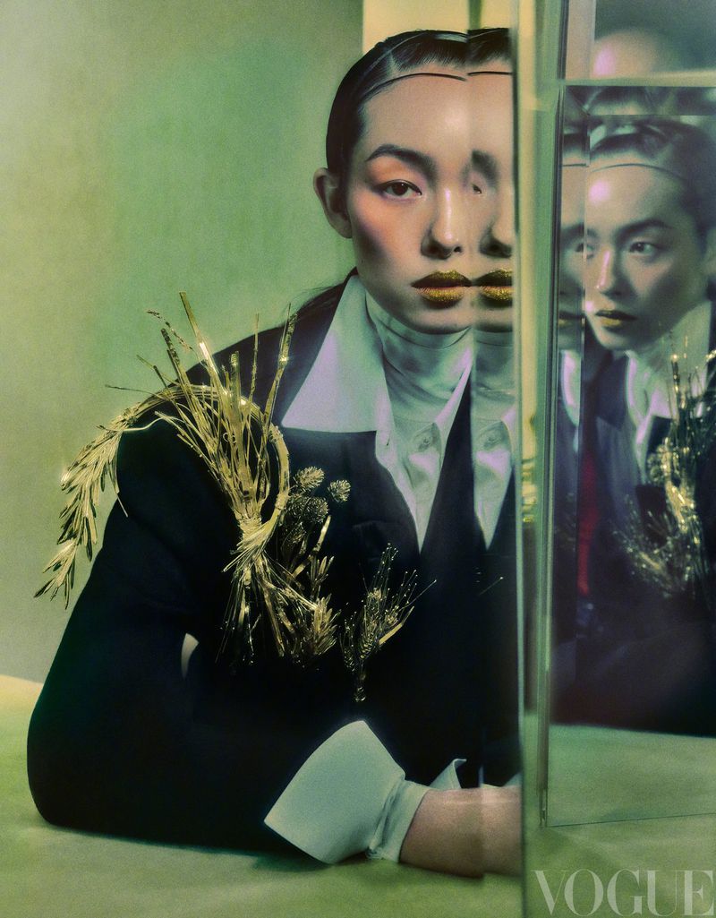 Fashion portrait of a woman in a black suit with gold embellishments, with a triple mirrored reflection, and green studio background. Vogue logo.