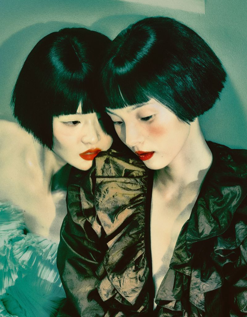 Two women with dark bob haircuts and red lipstick pose in front of a teal background. One wears a dark, ruffled top, and the other a light, ruffled top.