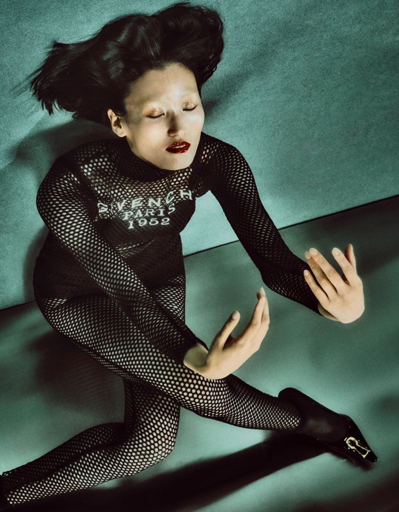 An Asian adult female poses in a black fishnet bodysuit and black heels against a teal backdrop with studio lighting and fashion styling.