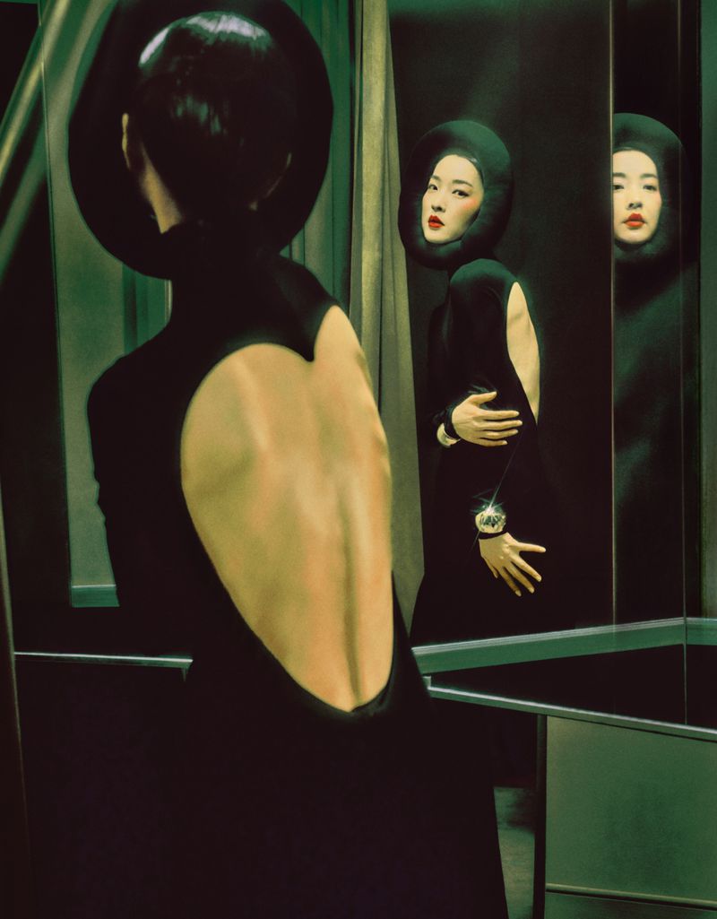 Woman in black dress with open back poses near mirror reflecting her image in an indoor setting with muted green tones.