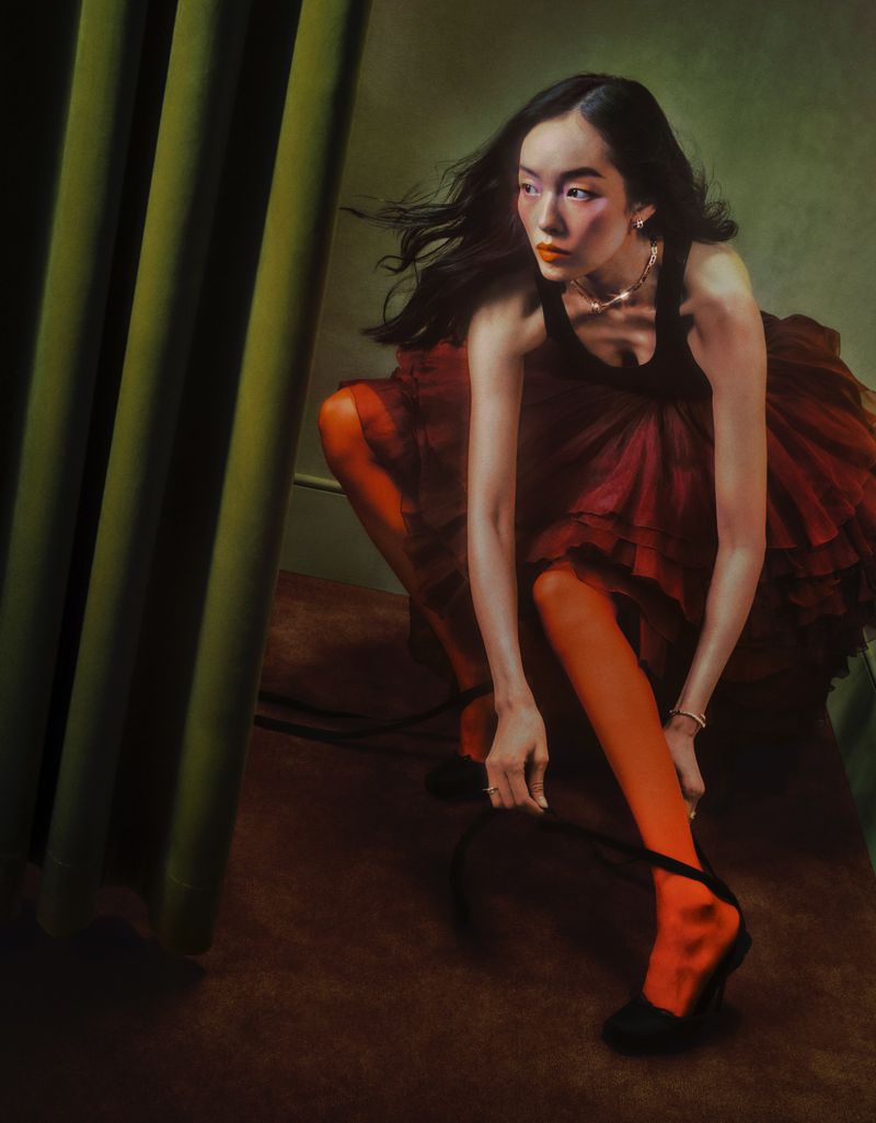 Elegant Asian woman in dark dress and orange tights kneels tying her black heels in an indoor setting near a green curtain.