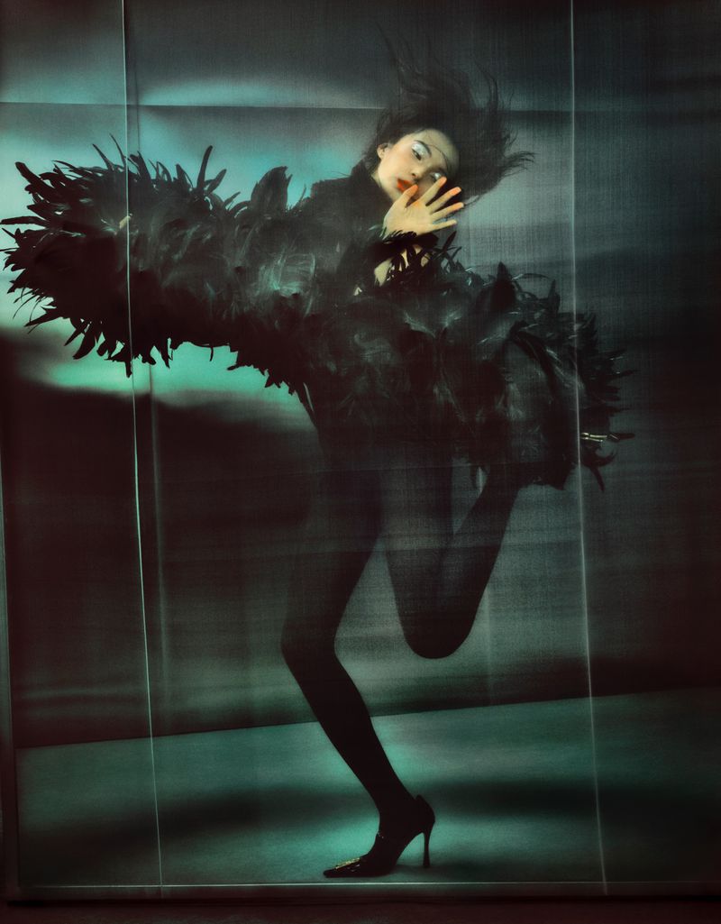 A woman with dark hair wearing a feathered black outfit poses in front of a glossy blue backdrop in this studio shot.