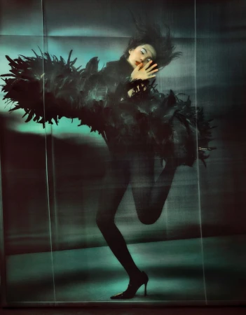 A woman with dark hair wearing a feathered black outfit poses in front of a glossy blue backdrop in this studio shot.