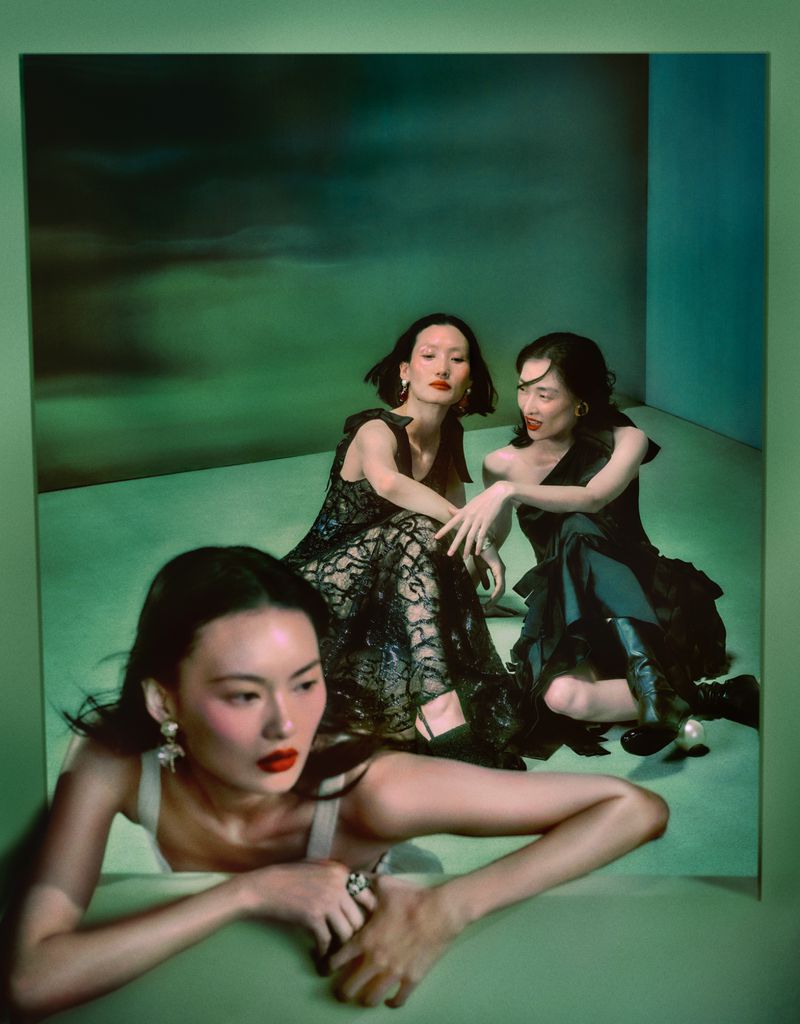 Three elegant women in black dresses pose in a studio with green lighting. The women wear elegant earrings and red lipstick.