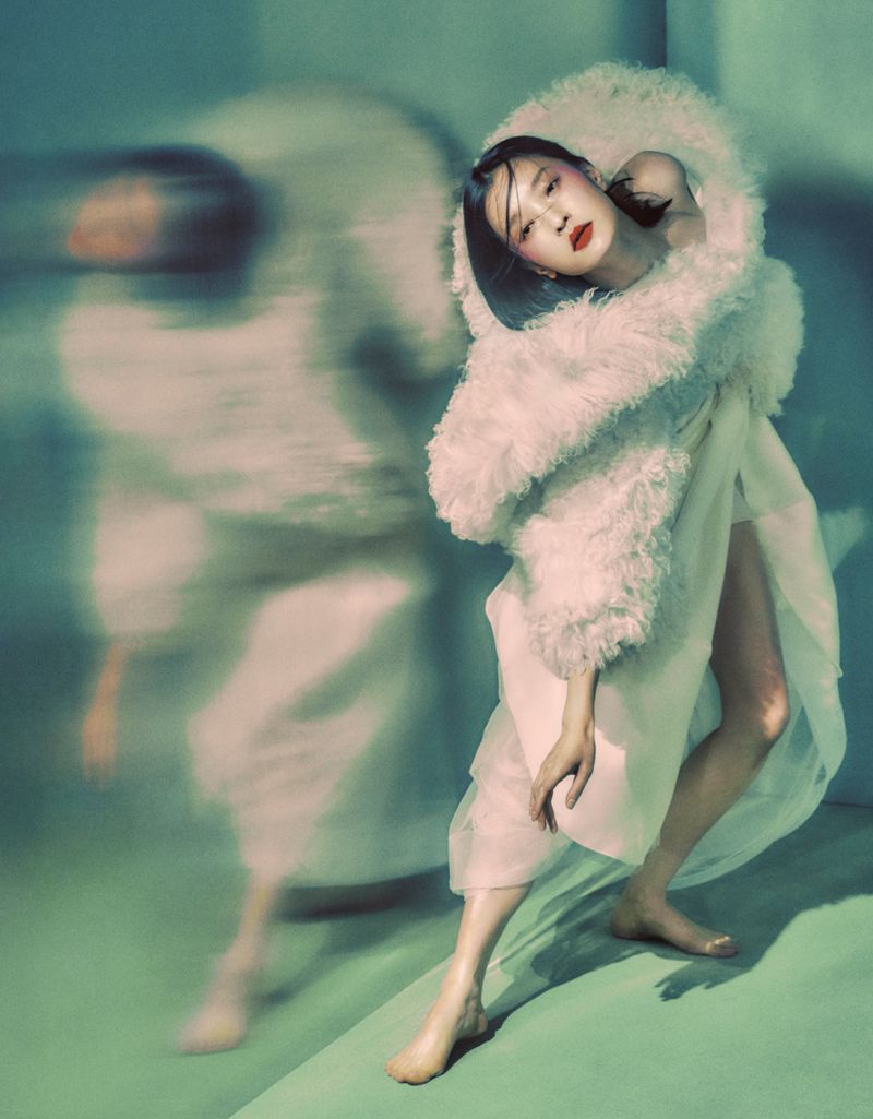 Fashion shot of a barefoot Asian woman in a draped white outfit, with a blurred motion effect figure, against a light blue background.