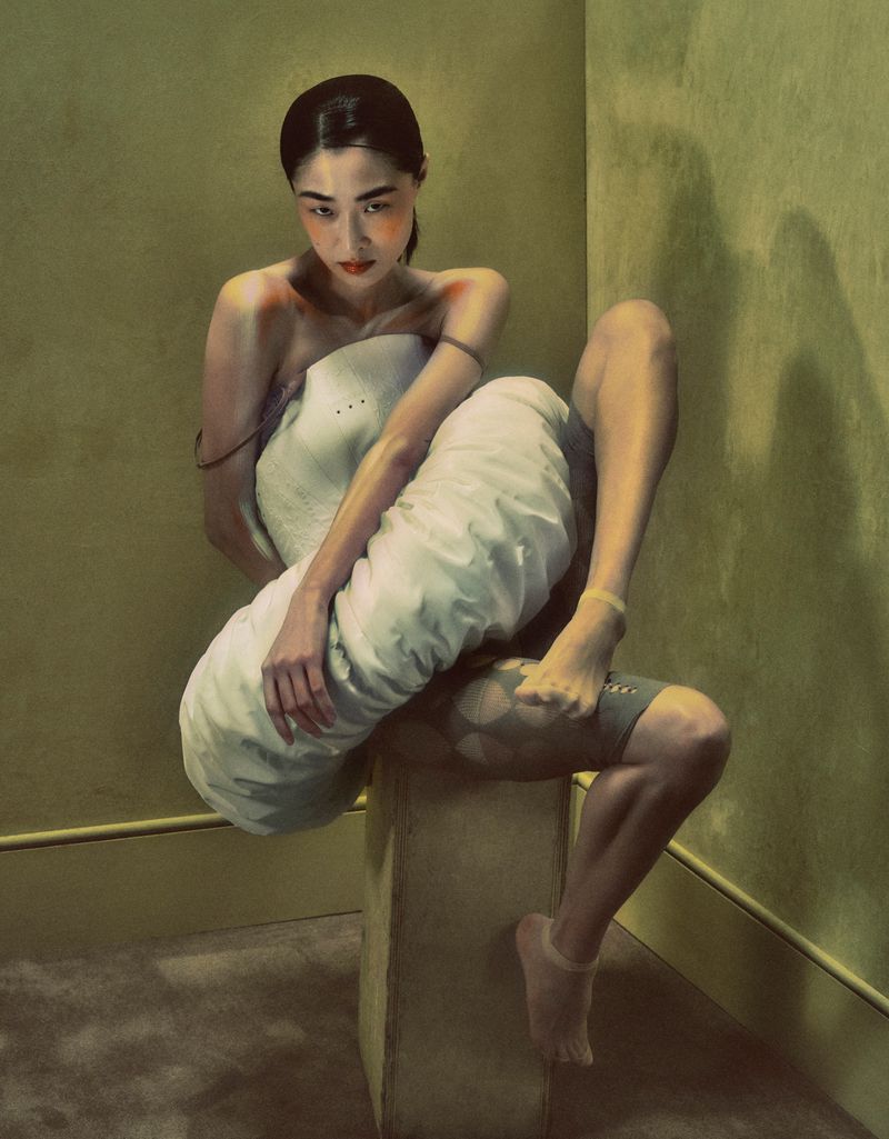 Digital painting features a woman sitting on a cube, wrapped in white fabric against a textured green wall, rendered in a painterly style.