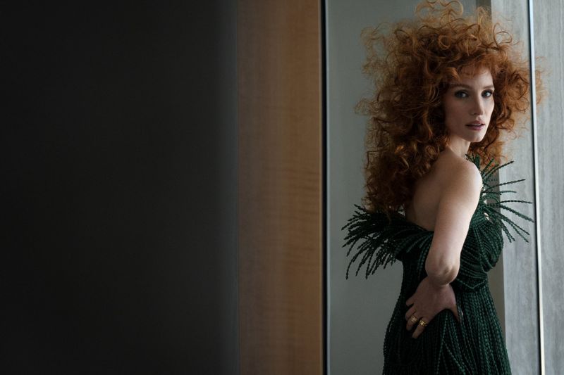 Woman with curly red hair, wearing a green textured dress, poses indoors near a paneled wall, looking over her shoulder at the camera.