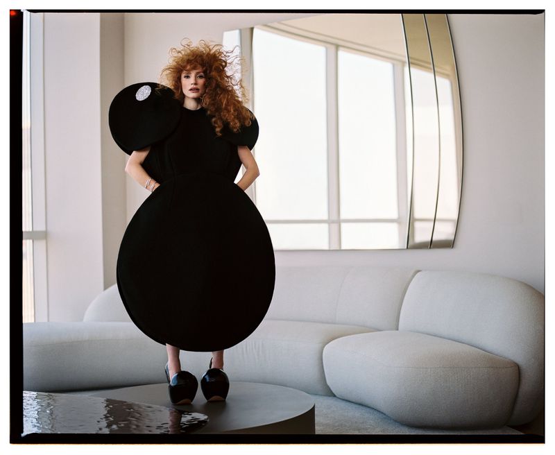 Fashionable woman with red curly hair poses in a striking black round dress and platform heels, inside a modern minimalist apartment.