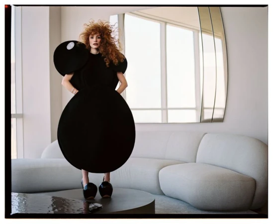 Fashionable woman with red curly hair poses in a striking black round dress and platform heels, inside a modern minimalist apartment.