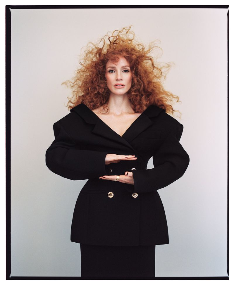 Portrait of an adult woman with red curly hair posing in a black double-breasted blazer and skirt against a neutral background.