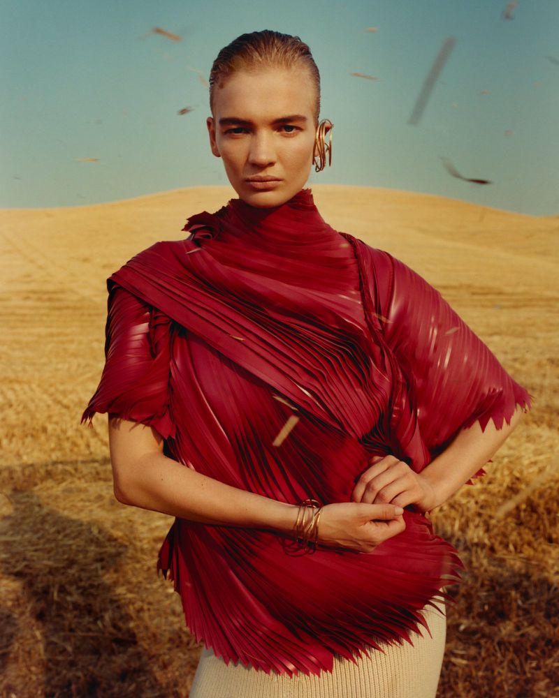 Woman in pleated burgundy top poses in arid field under blue sky with gold ear cuffs and bracelets, creating a fashion editorial.