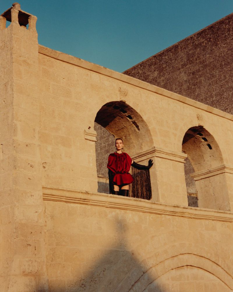 A woman in a red dress and black gloves stands on a stone balcony with arched windows, against a background of a building and blue sky.