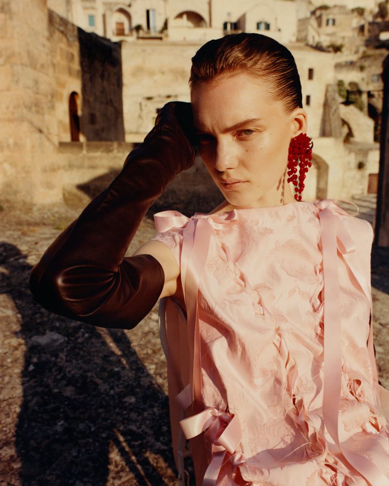 Portrait of a woman in a pink dress and long gloves posing in an outdoor setting with stone buildings in the background on a sunny day.