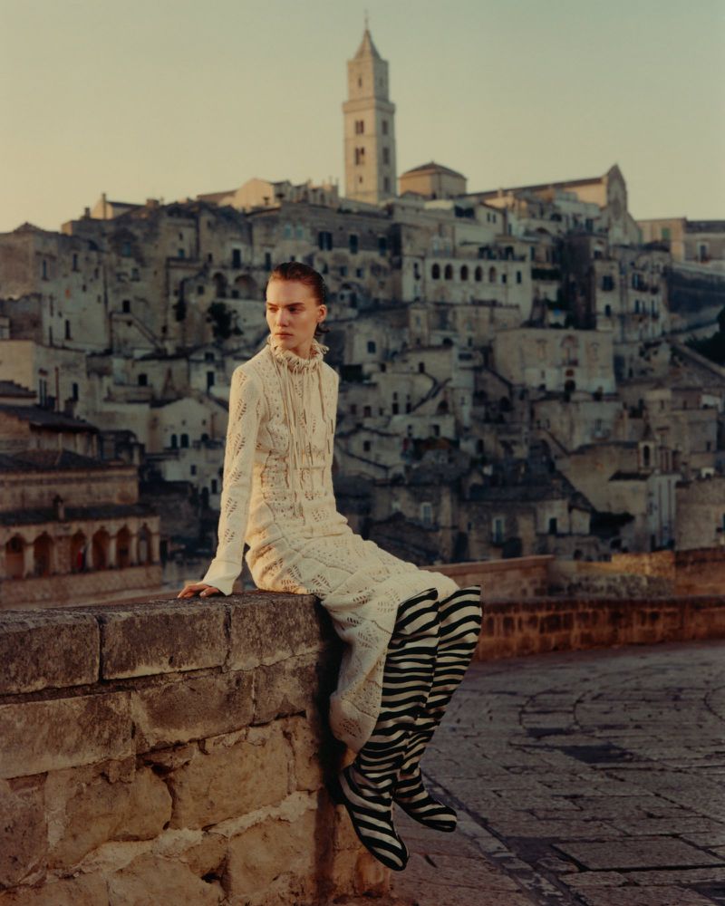Fashion editorial shot of a young woman in a textured dress and striped boots, sitting on a stone wall in an old European town.
