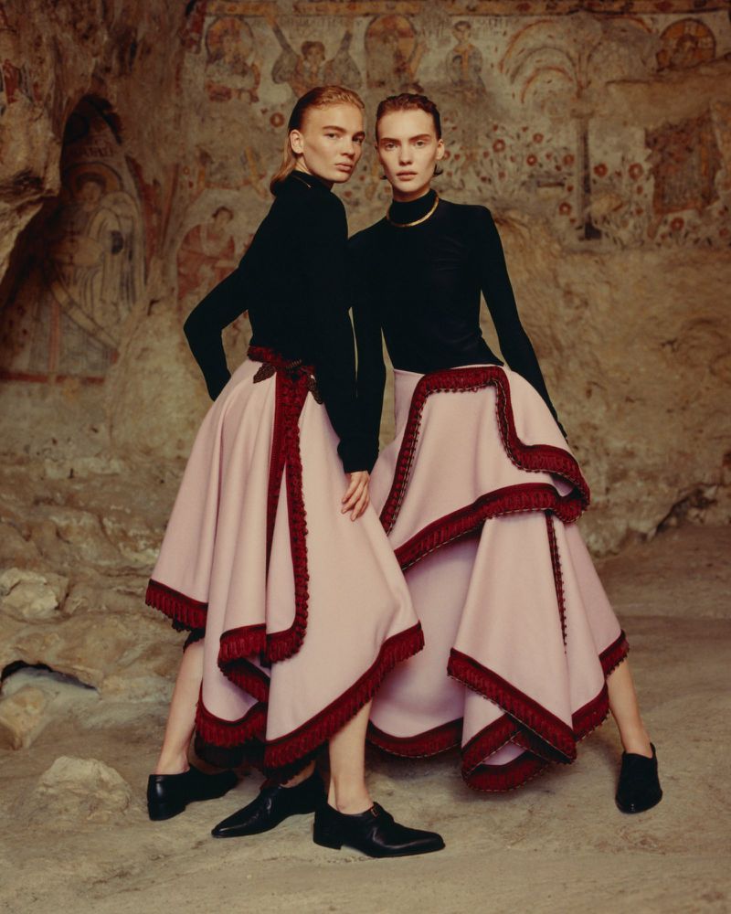 Two fashion models pose in black tops and tiered pink skirts with red trim in front of an aged wall with faded paintings.