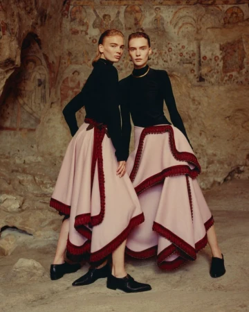 Two fashion models pose in black tops and tiered pink skirts with red trim in front of an aged wall with faded paintings.