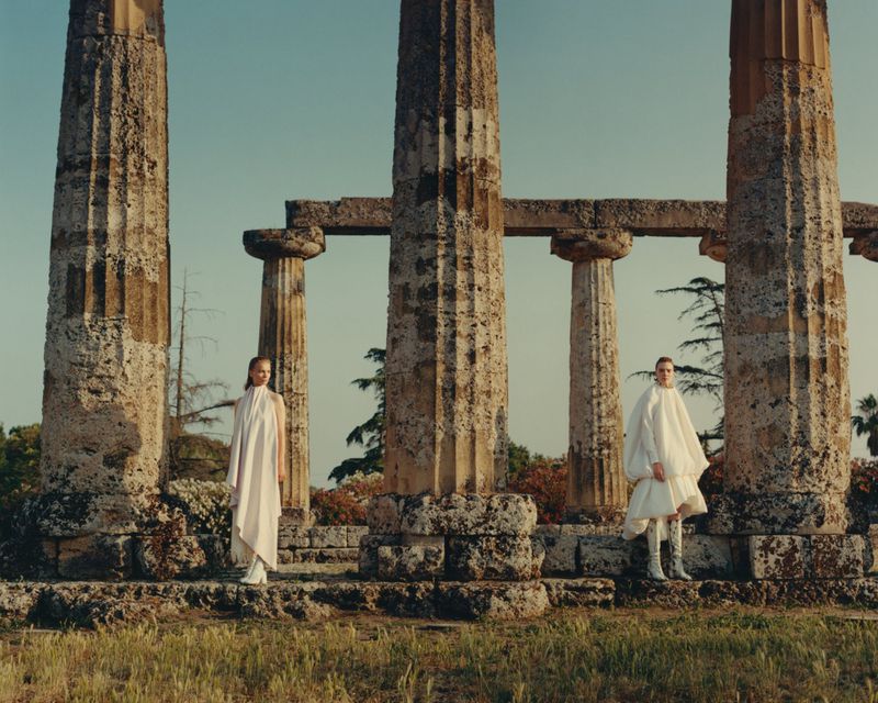 Two fashion models in white garments stand between stone columns against a blue sky in an outdoor setting on a sunny day.