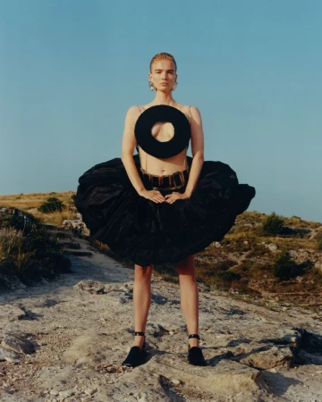 Model wearing a sculptural black cutout circular top and voluminous puffed skirt on a rocky terrain under a clear sky.