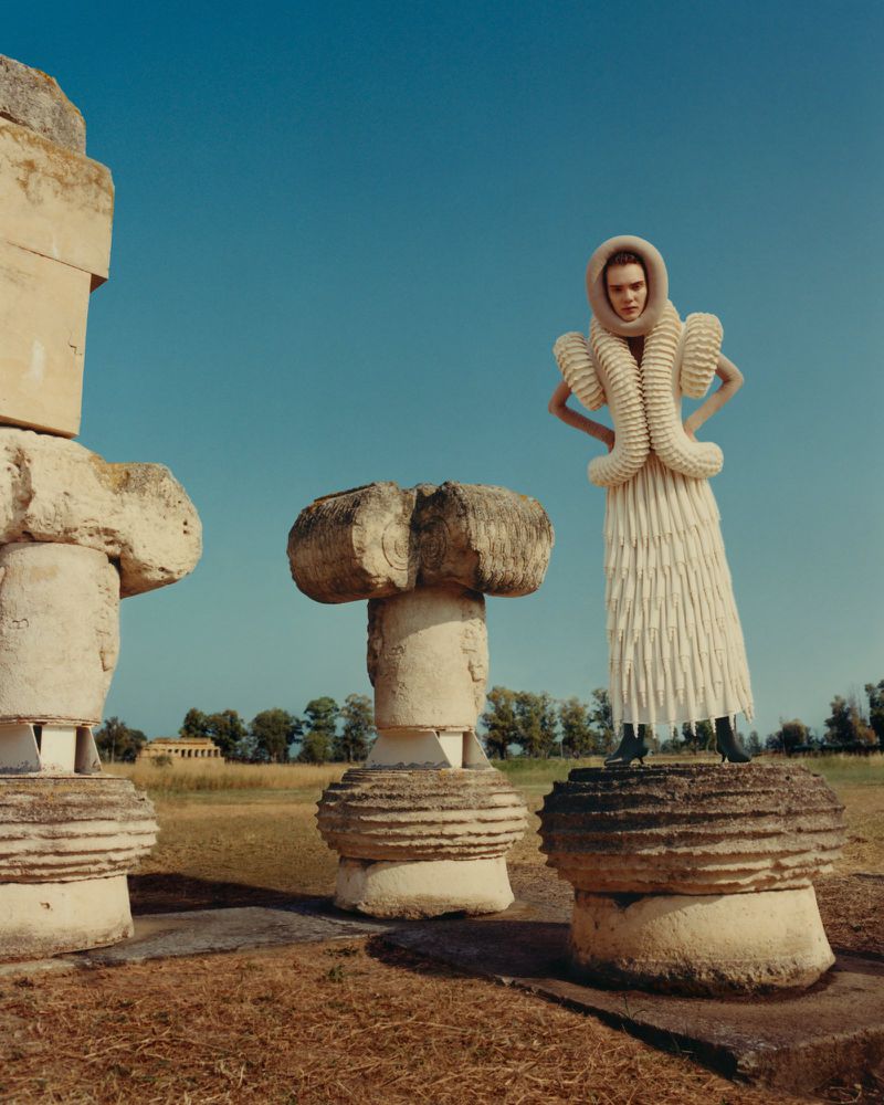 A woman stands on a stone pedestal in a cream-colored gown with a unique collar with stone ruins in the background on a clear day.