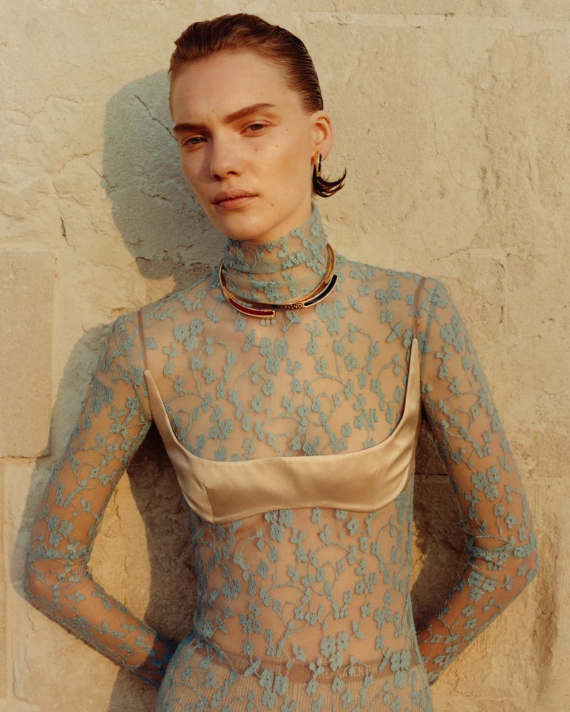 A fair-skinned woman with wet-look hair poses in a high-necked, floral-patterned lace dress with a light peach-colored bralette underneath.