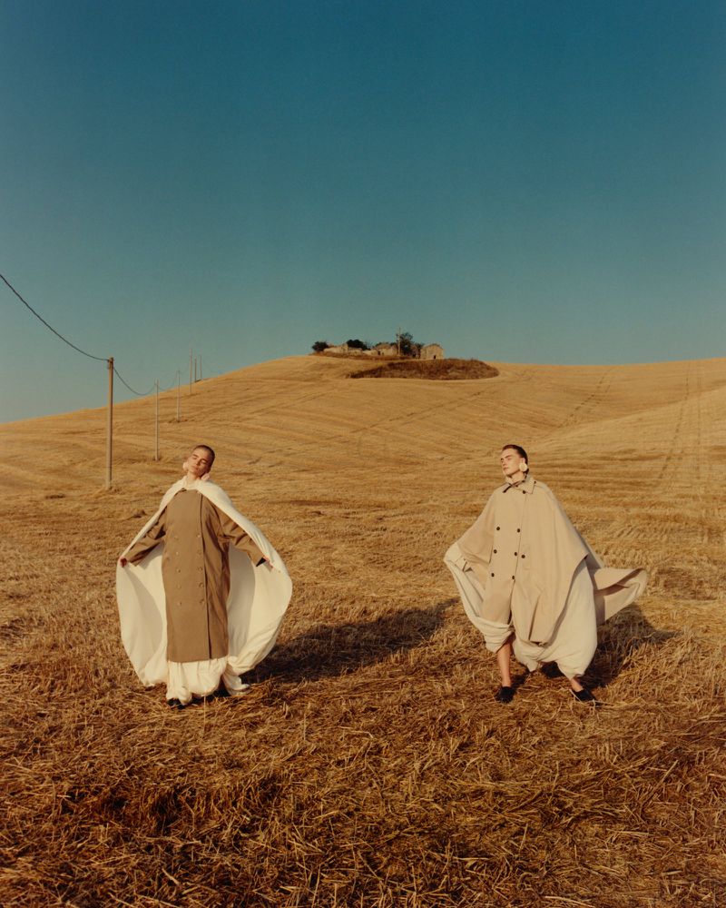 Two women in billowing neutral-toned outfits pose in a golden field under a clear blue sky on a sunny day, evoking a fashion editorial.