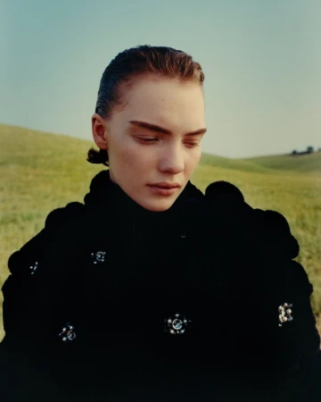 Model looking downward wearing a black garment adorned with jewel brooches against rolling green hills.