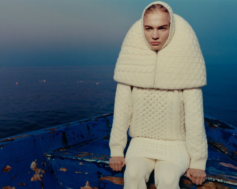 Fashion shot of androgynous model with neutral expression wearing a thick cream-colored knit sweater and hooded shawl sitting on a blue boat.