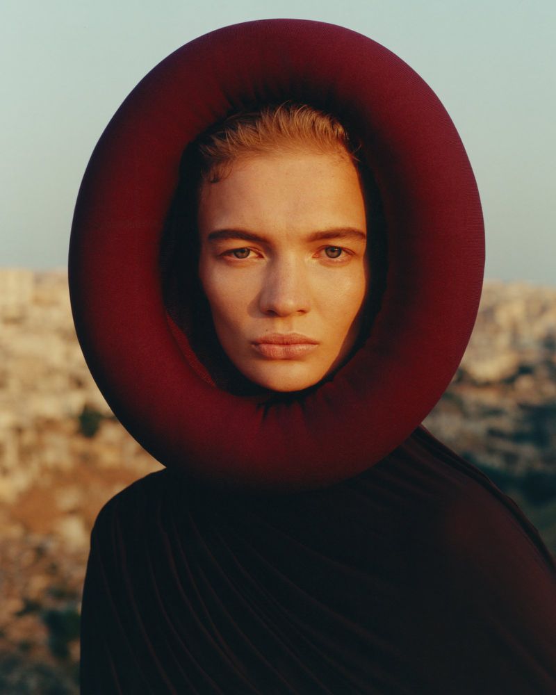 Woman in dark garment and red circular headpiece poses outdoors with blurred background in eye-level shot, medium framing, and warm lighting.