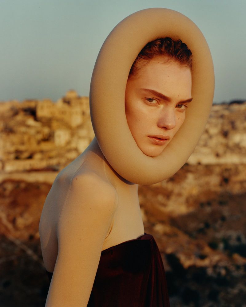 Elegant portrait of a fair-skinned woman wearing a tan circular collar piece, posing in front of a blurred arid, mountainous landscape.