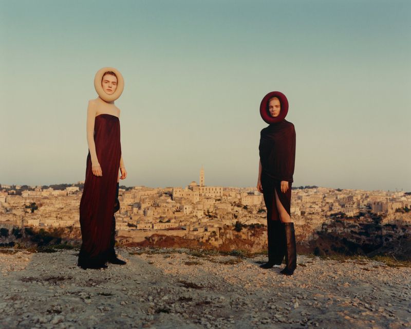 Two women in long dresses and headwear stand on a hilltop overlooking the town of Matera, Italy, under a clear sky during the day.