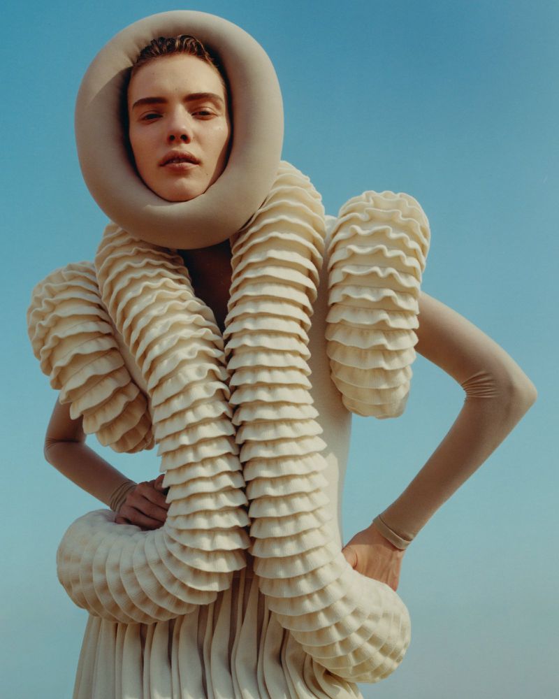 Fashion shot of a person wearing a sculptural cream dress with tube accents, hands on waist, against a blue sky background.