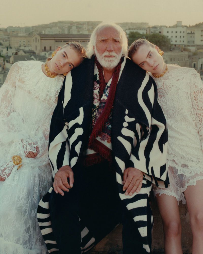 An elderly man with a white beard poses between two young women with blonde hair wearing white lace dresses against a city background.